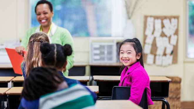 An image of an Asian American girl smiling at the camera in a diverse classroom. Credit: Canva.